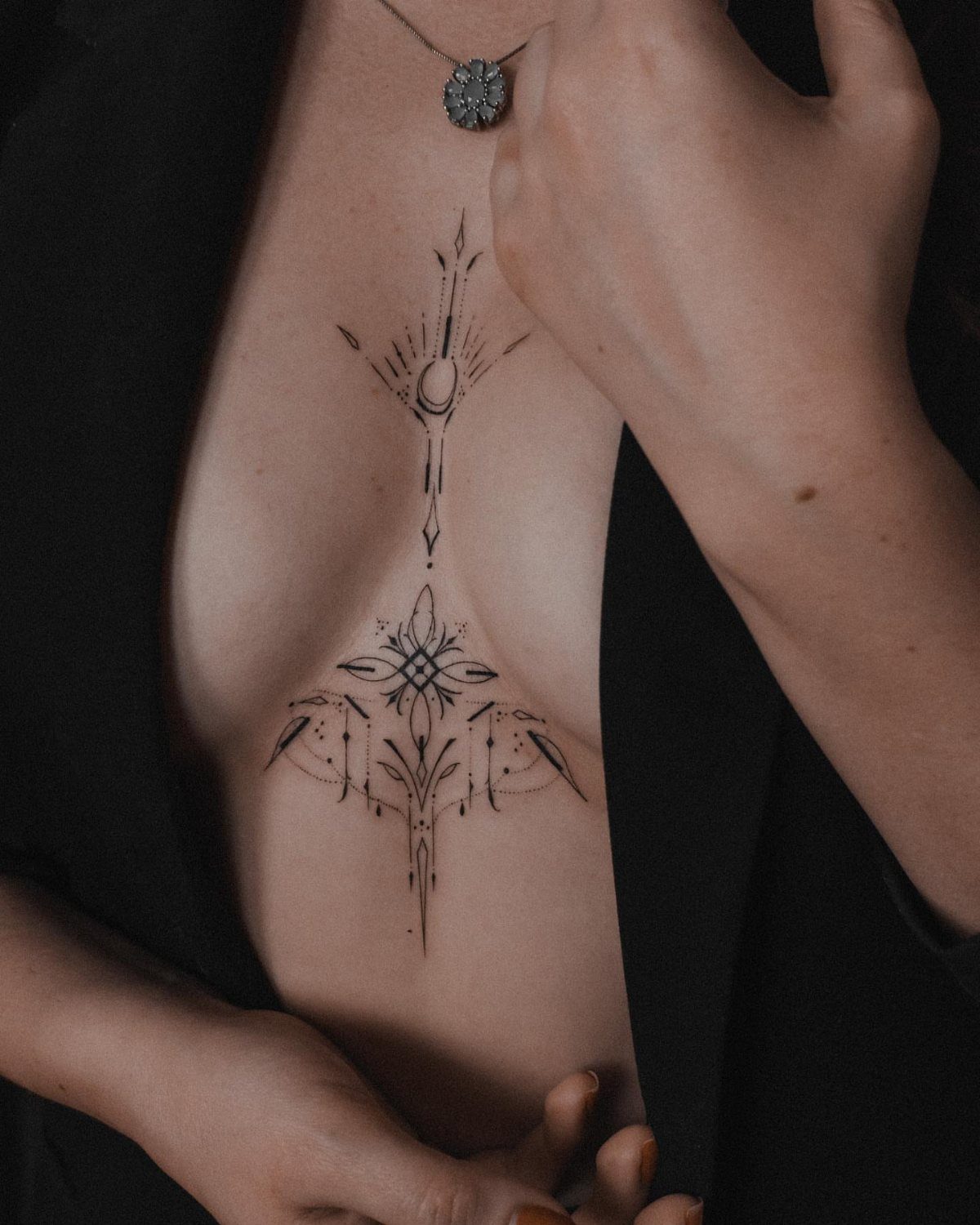 9 Chest Tattoo Ideas for Women: Floral, Sternum, Underboob & More 2026 7 A woman's chest featuring a symmetrical black ink tattoo with fine geometric lines and abstract shapes.