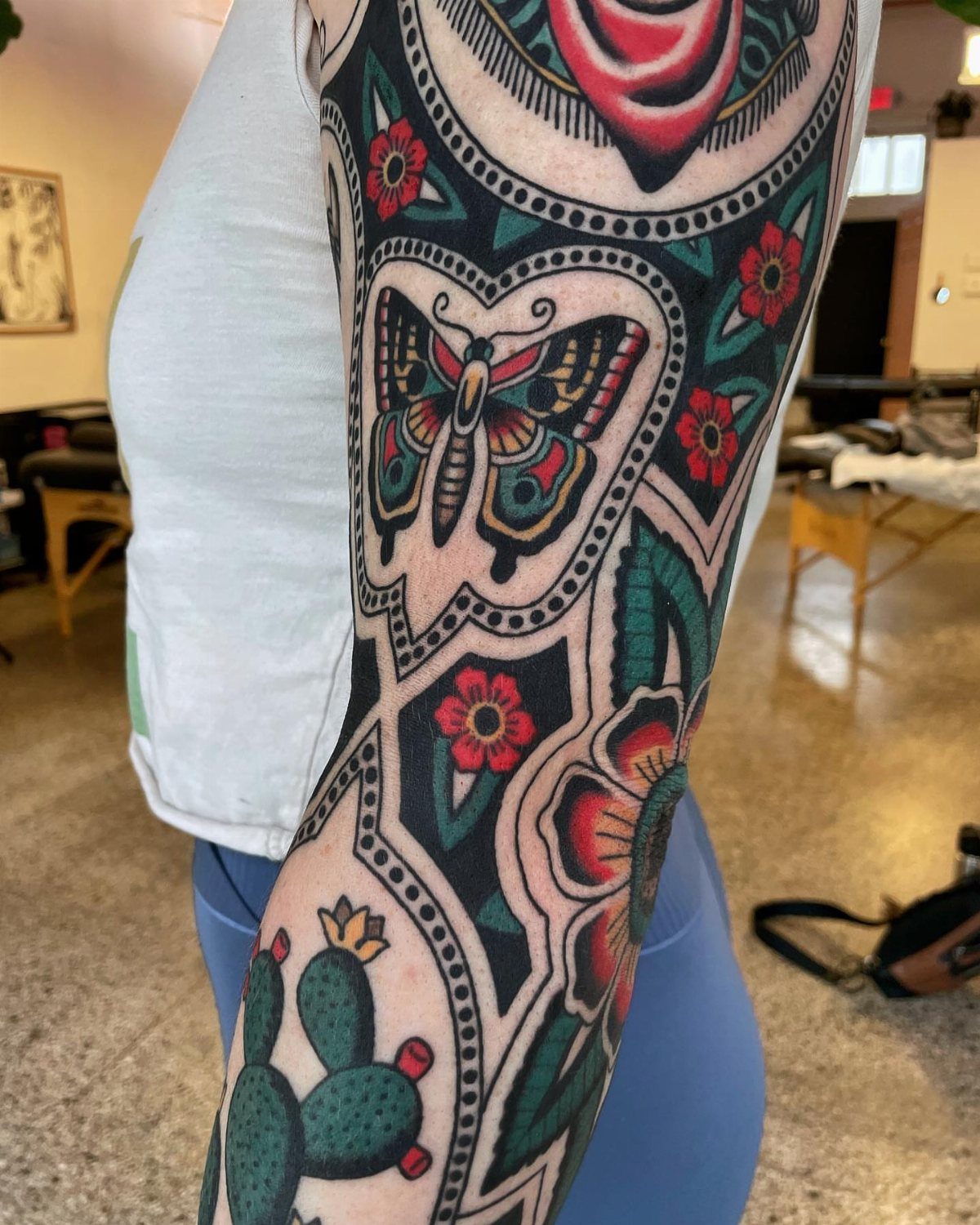 Arm covered in colorful American traditional tattoos featuring a butterfly, red flowers, and a green cactus.