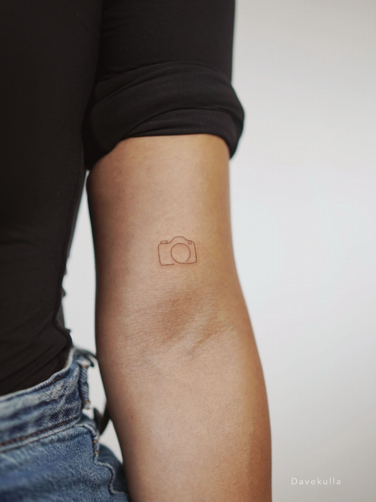 Fine Line Travel Tattoo: 10 Designs That Stay Light on Skin 7 A small fine line camera tattoo on a person's upper arm against a white background.