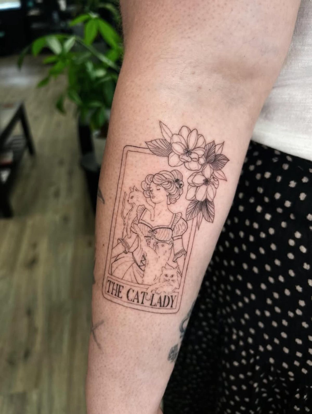 A fine line black ink tattoo of a tarot card titled The Cat Lady on a forearm.