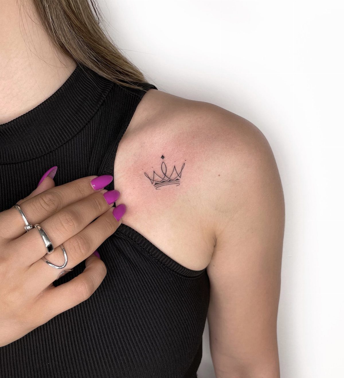 A small fine-line crown tattoo on a person's shoulder next to purple fingernails.