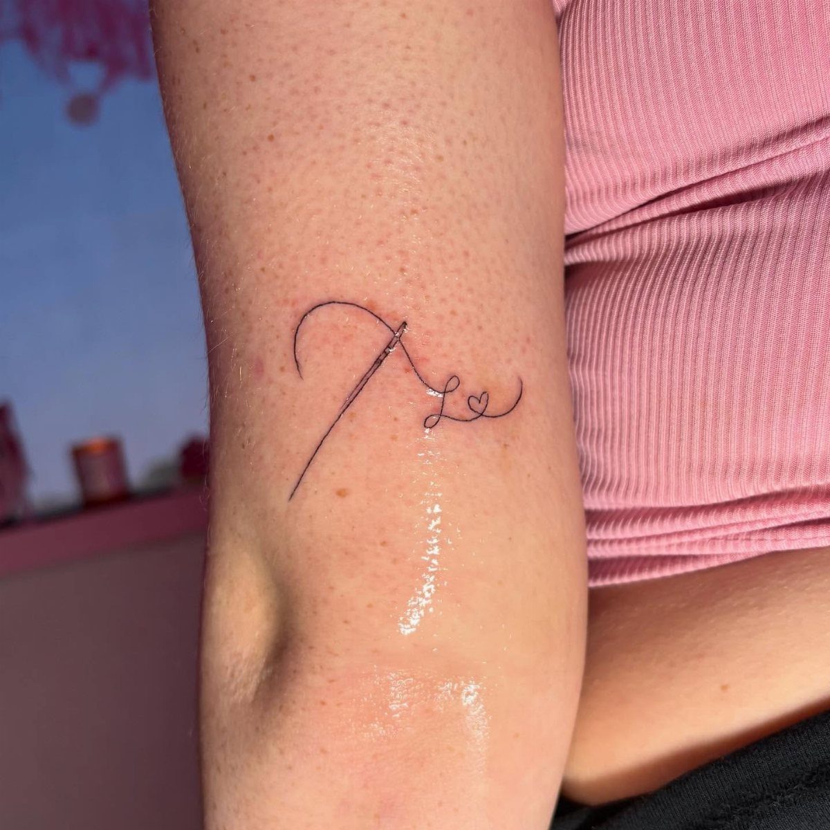 A small minimalist tattoo of a needle and thread forming a heart on an arm.