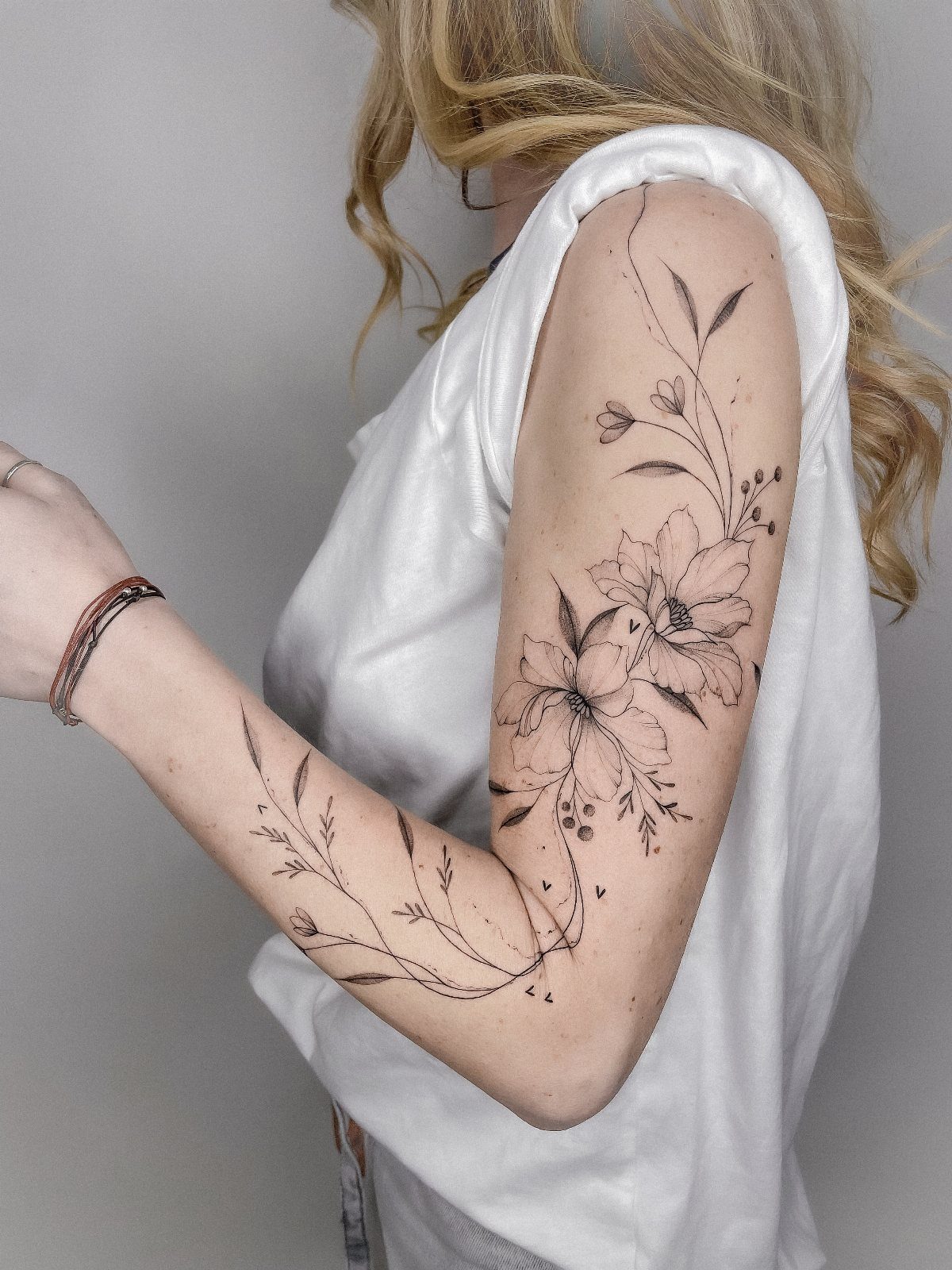 A person's arm featuring a delicate black ink floral tattoo with fine lines and leaves.