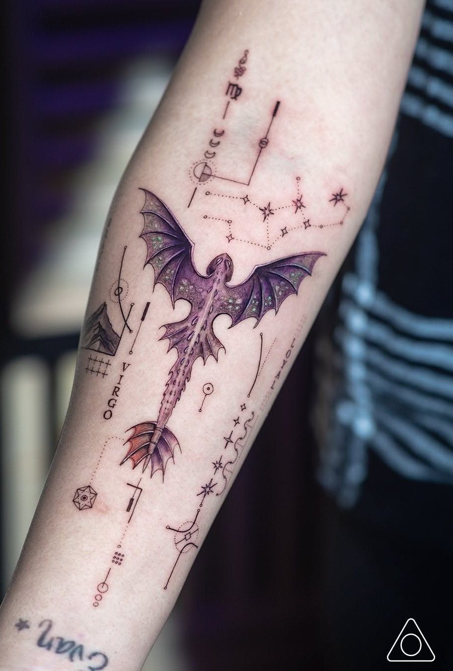 A purple dragon tattoo on a forearm surrounded by delicate black ink celestial symbols and constellations.