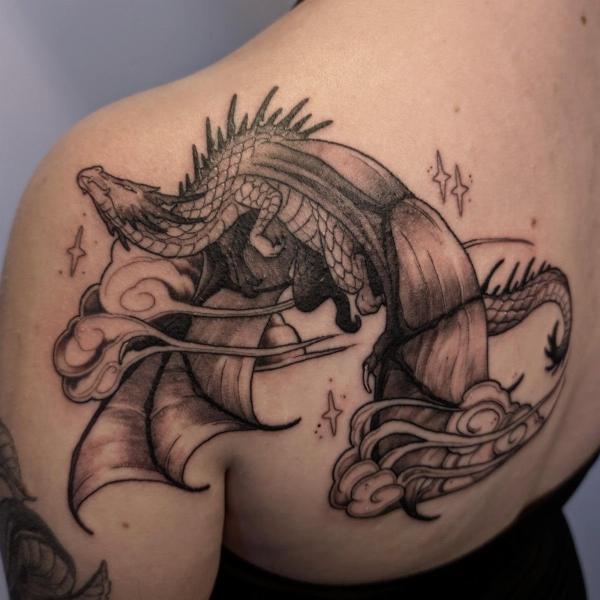 6 Dragon Backpiece Tattoo Ideas for Your Next Session 6 A black and grey dragon tattoo with large wings winding through clouds on a person's skin.