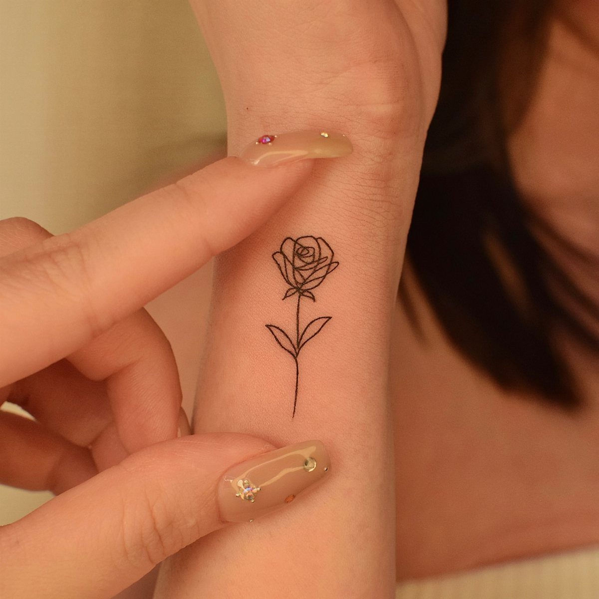 A fine line rose tattoo on a person's wrist with manicured nails.