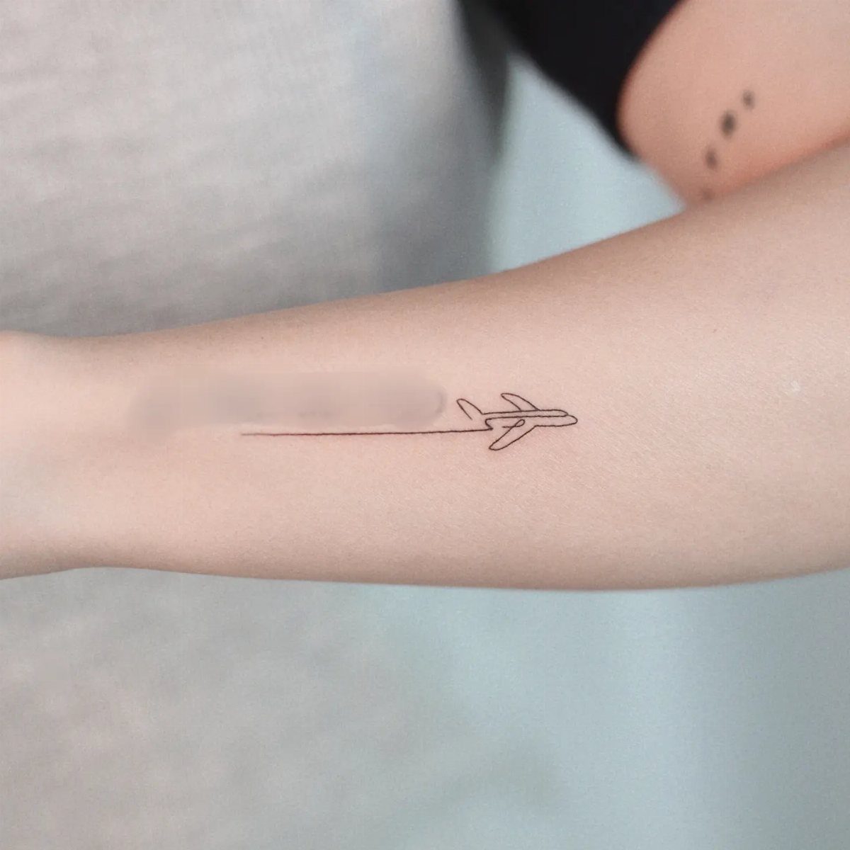 A minimalist fine line tattoo of an airplane silhouette on a person's forearm.