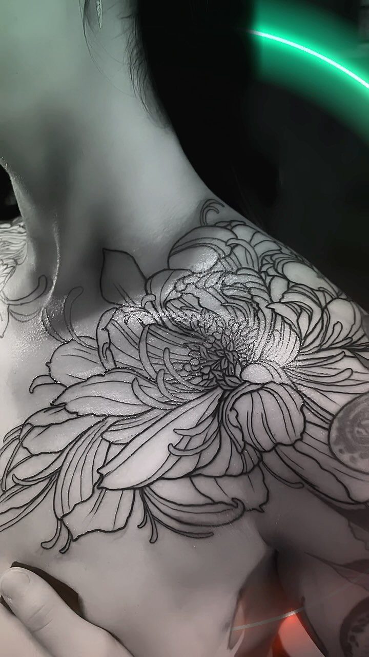9 Chest Tattoo Ideas for Women: Floral, Sternum, Underboob & More 2026 2 Large black ink chrysanthemum tattoo covering a woman's upper chest and shoulder area.