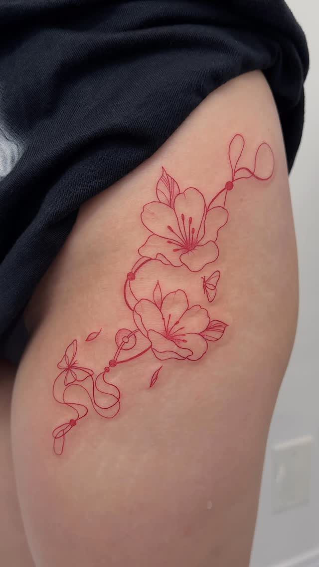 Thigh Tattoos for Women: 8 Ideas from Floral to Geometric Art 2 A red fine-line tattoo of two flowers and butterflies on a person's thigh.