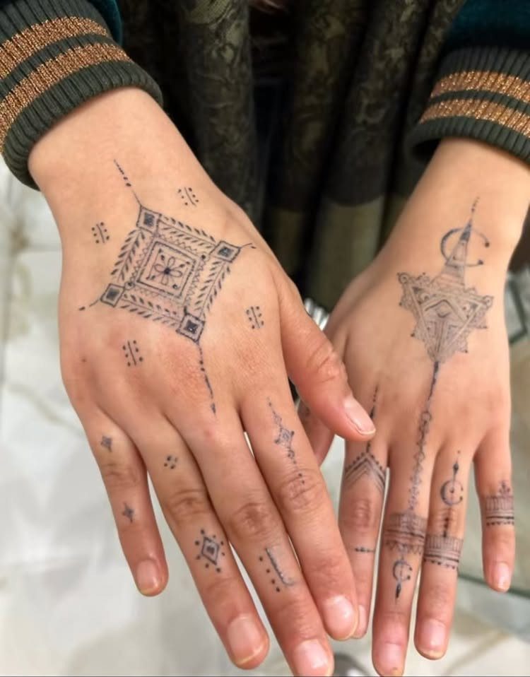 Two hands featuring intricate black henna tattoos with various geometric patterns and diamond shapes.