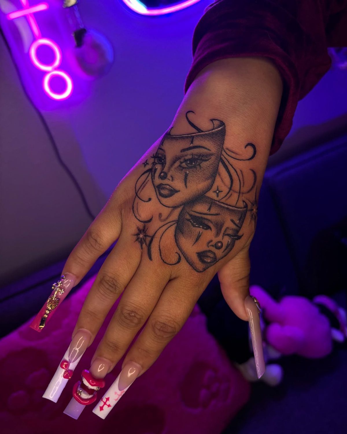 A hand with long decorative nails displaying a black ink tattoo of two theatrical masks.