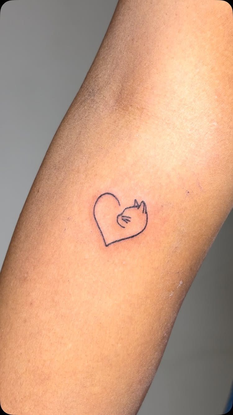 A small fine line tattoo of a heart shape containing a cat silhouette on skin.