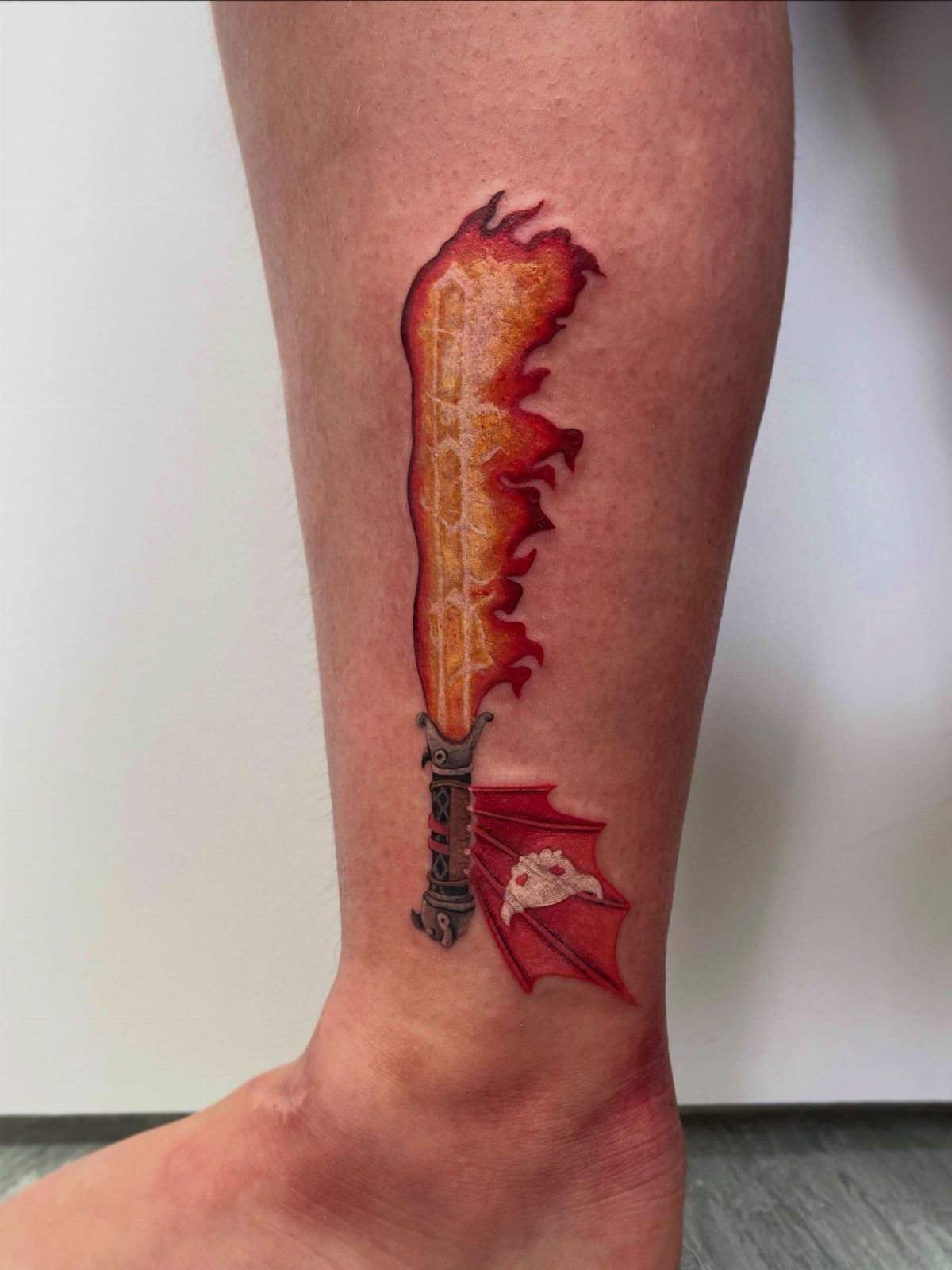 A detailed tattoo of a flaming sword with a dragon wing on a person's leg.
