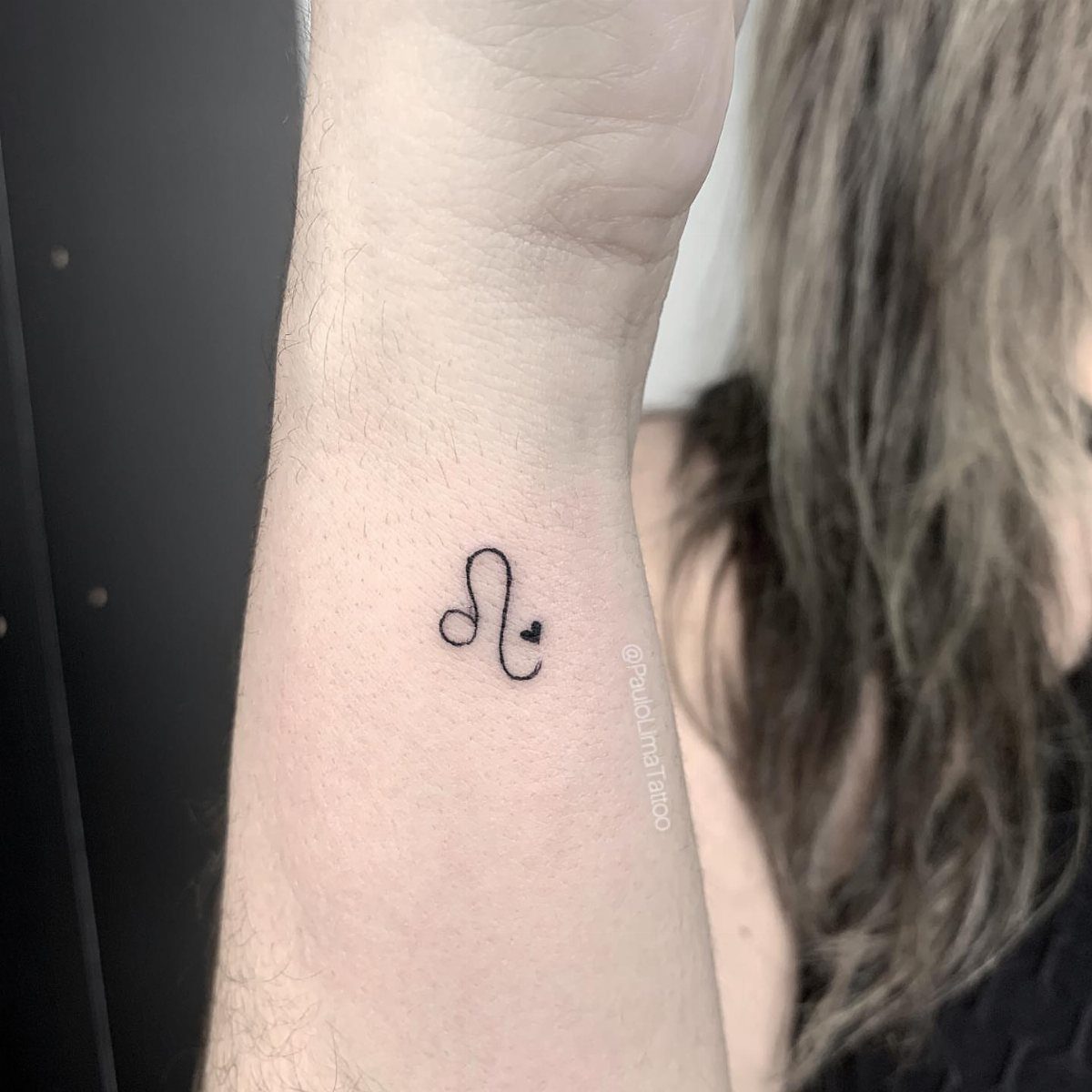 A small black ink Leo glyph tattoo with a heart detail on a person's wrist.