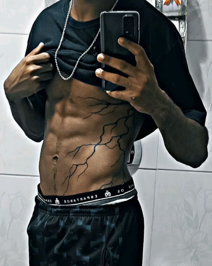 A man showing a large black lightning bolt tattoo on his abdomen in a mirror selfie.
