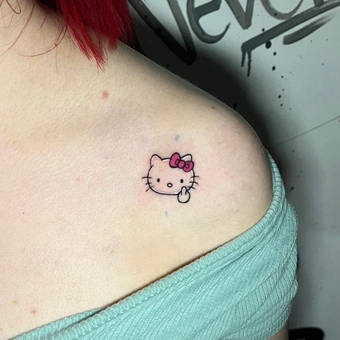 A tiny Hello Kitty face tattoo with a pink bow on a person's shoulder.