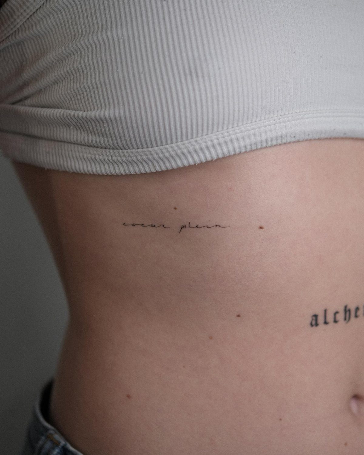 Small minimalist script tattoos on a person's side ribs in fine black ink.
