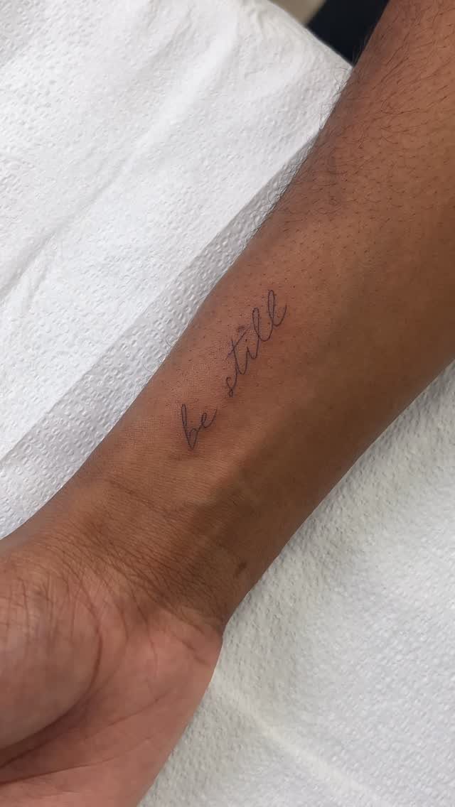 22 Delicate Tattoo Ideas for Light, Fine-Line Ink 14 A small script tattoo reading 'be still' on a person's forearm against a white background.