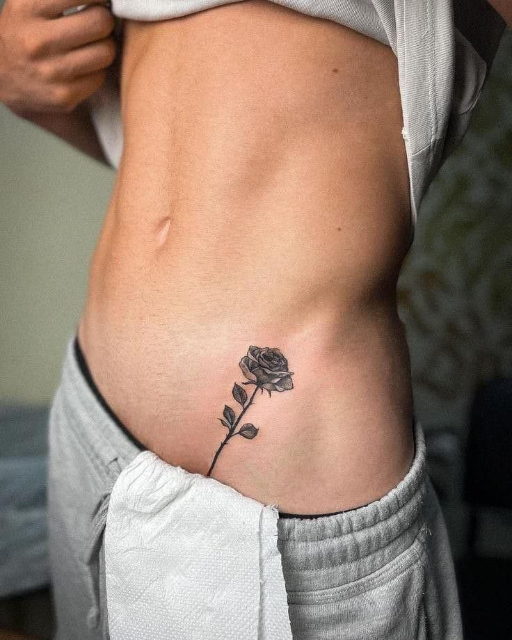 A realistic black and grey rose tattoo with a stem on a person's waist.