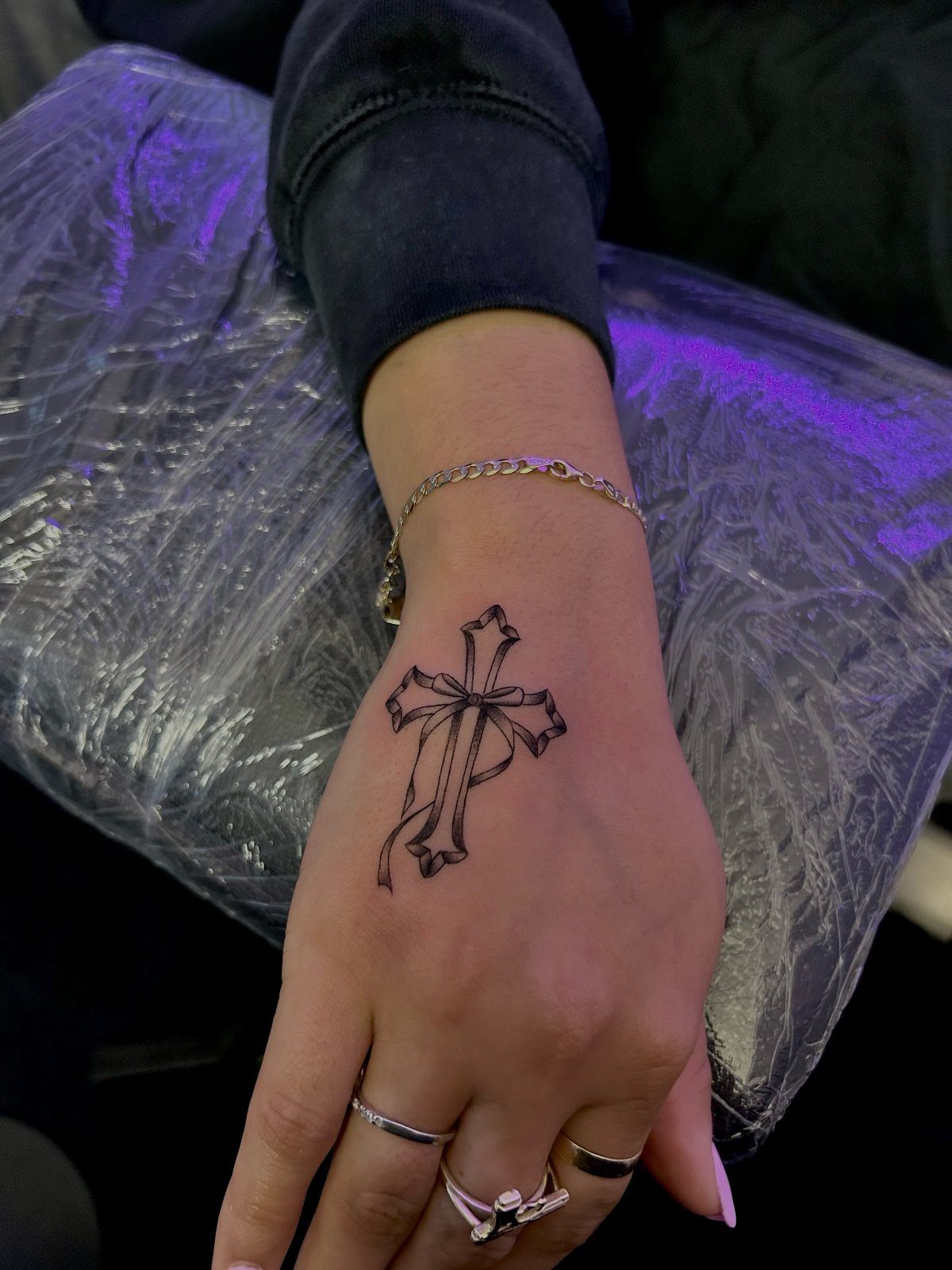 A hand with a small black cross tattoo and ribbon design, wearing silver rings.