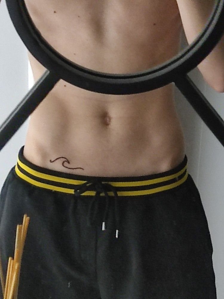 A man's abdomen featuring a small black single-line wave tattoo above his waistband.