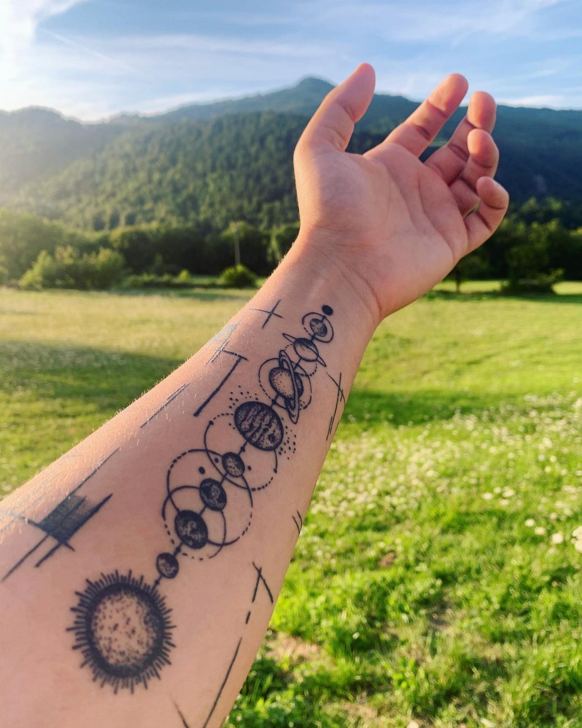 25 Space Themed Tattoos from Watercolor Cats to Black Holes 10 A forearm tattoo of the solar system with planets and a sun against a natural landscape.