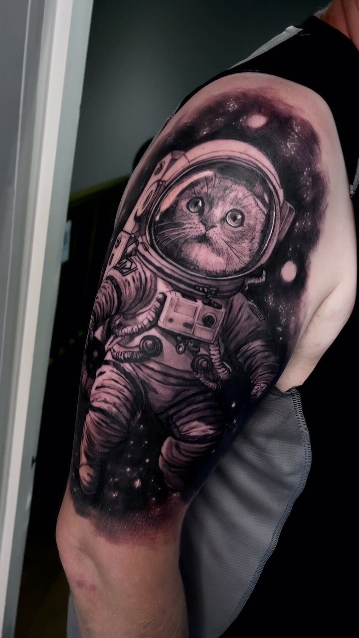 25 Space Themed Tattoos from Watercolor Cats to Black Holes 7 A detailed black and grey tattoo of a cat in an astronaut suit on a person's shoulder.