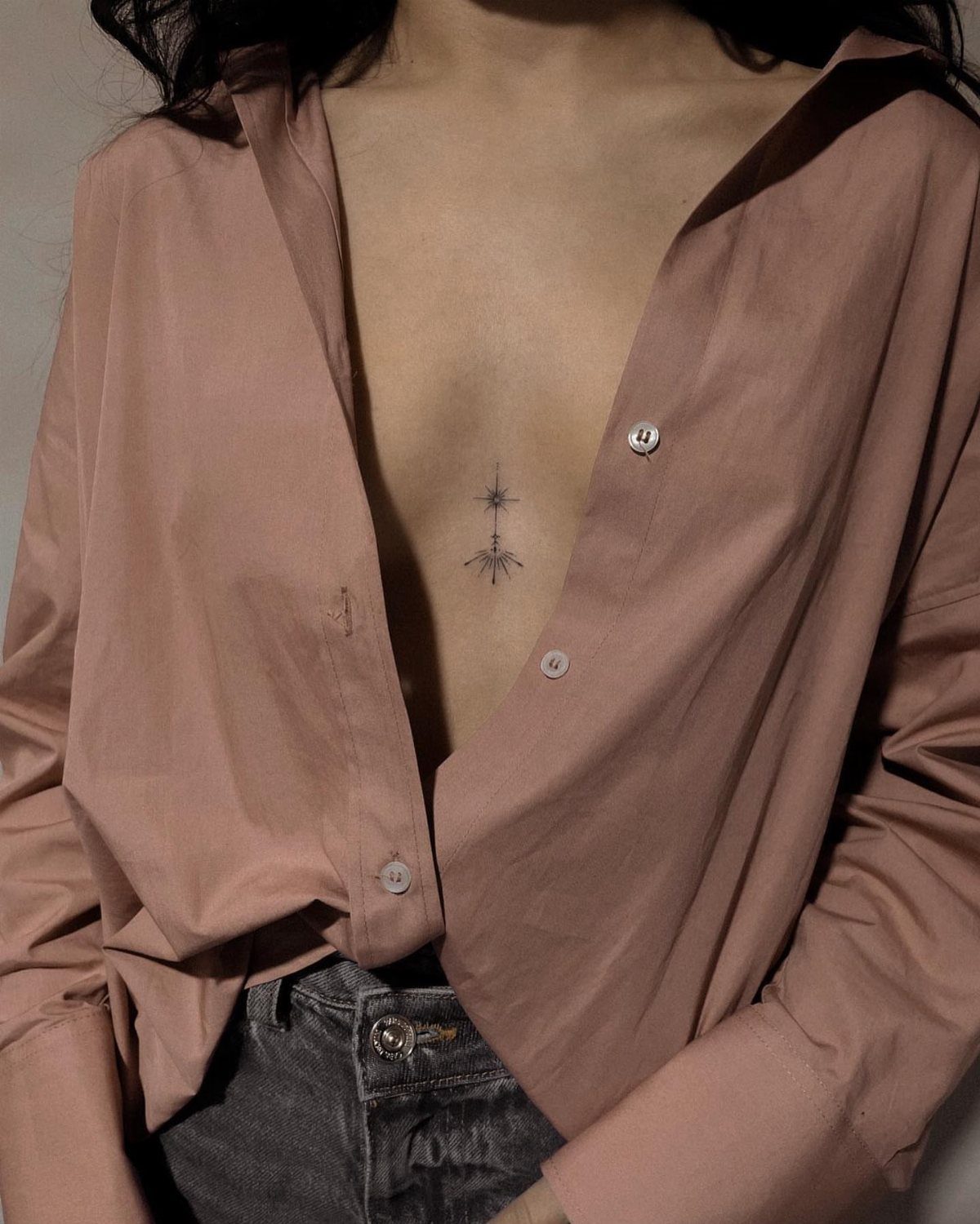 9 Chest Tattoo Ideas for Women: Floral, Sternum, Underboob & More 2026 3 A woman's sternum featuring a small, vertical black ink star tattoo under an open shirt.