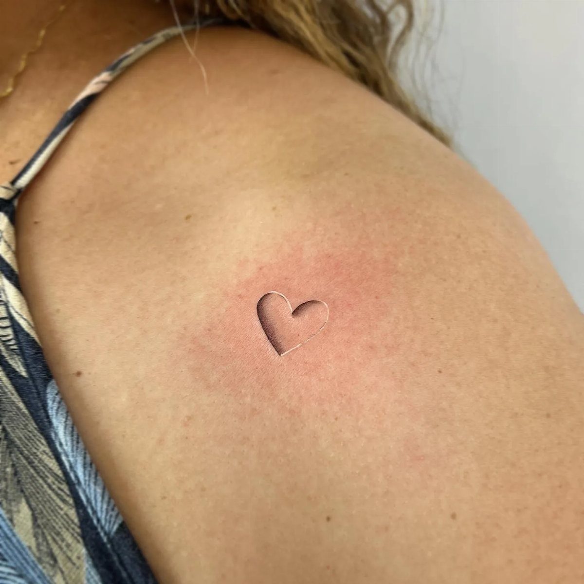 A small heart outline tattoo located on a person's shoulder skin.