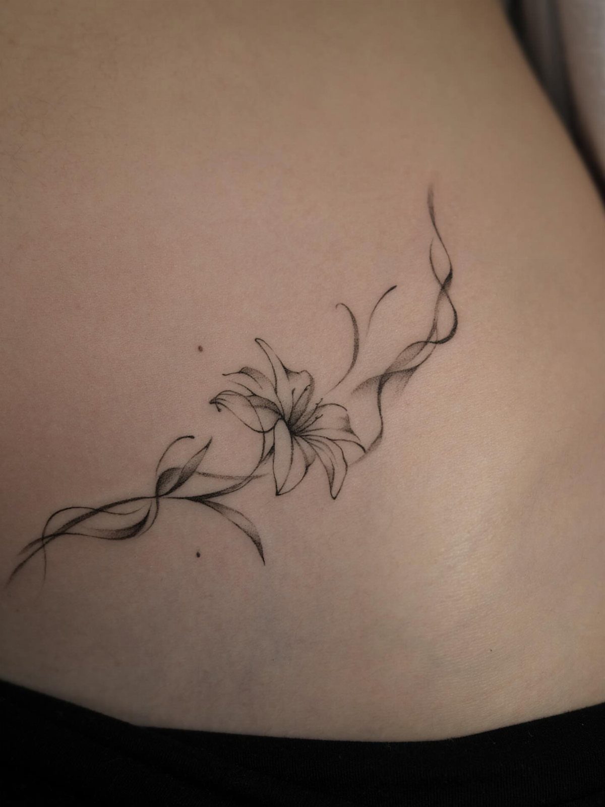 A fine line black ink lily tattoo on a person's lower stomach and hip.