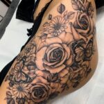 A detailed black and grey floral tattoo with roses and butterflies on a woman's thigh.