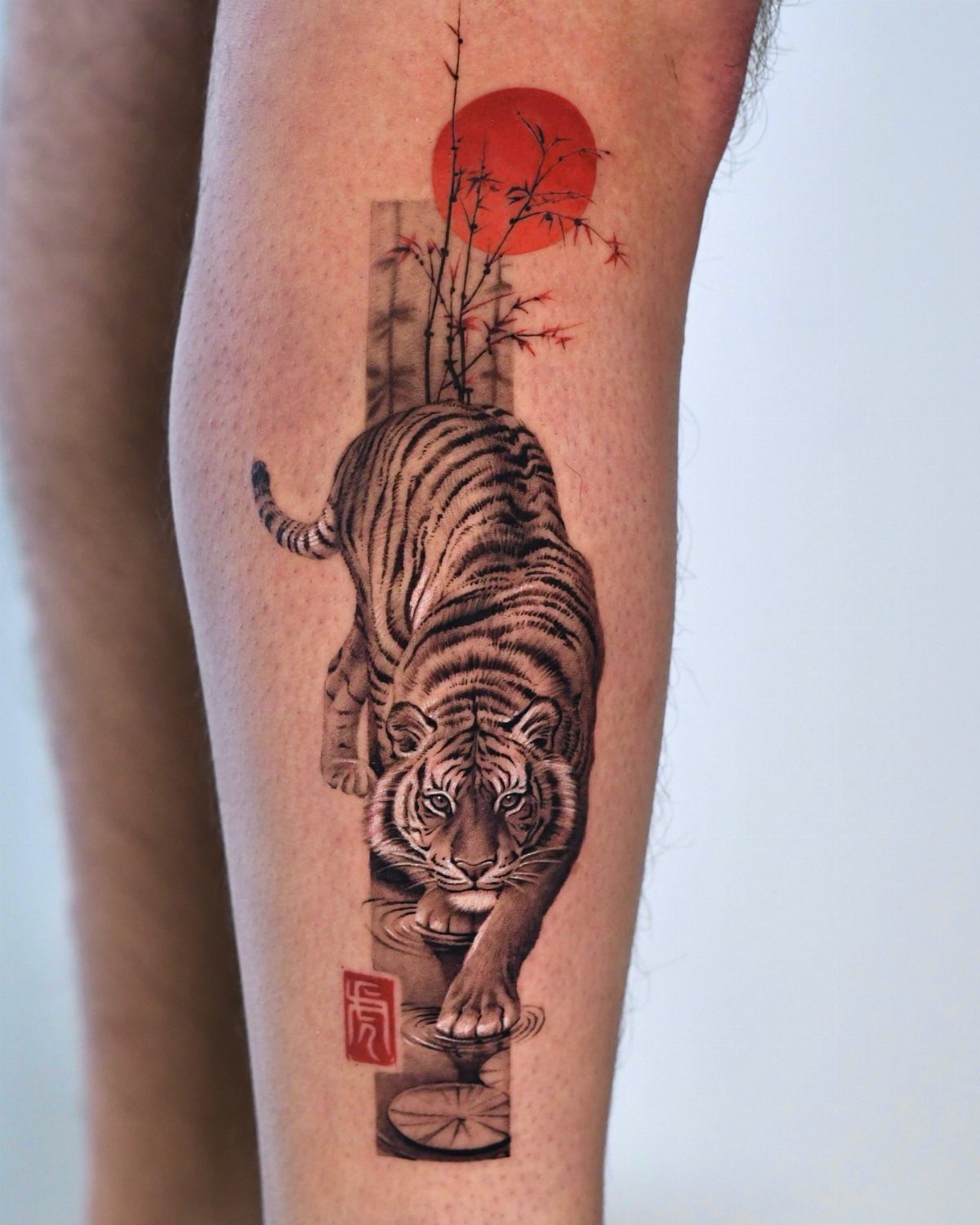 A detailed black and grey tiger tattoo with a red sun and bamboo on a person's leg.
