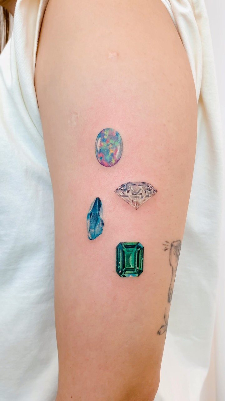 22 Delicate Tattoo Ideas for Light, Fine-Line Ink 16 Three colorful gemstone tattoos on a person's upper arm including an opal and emerald.