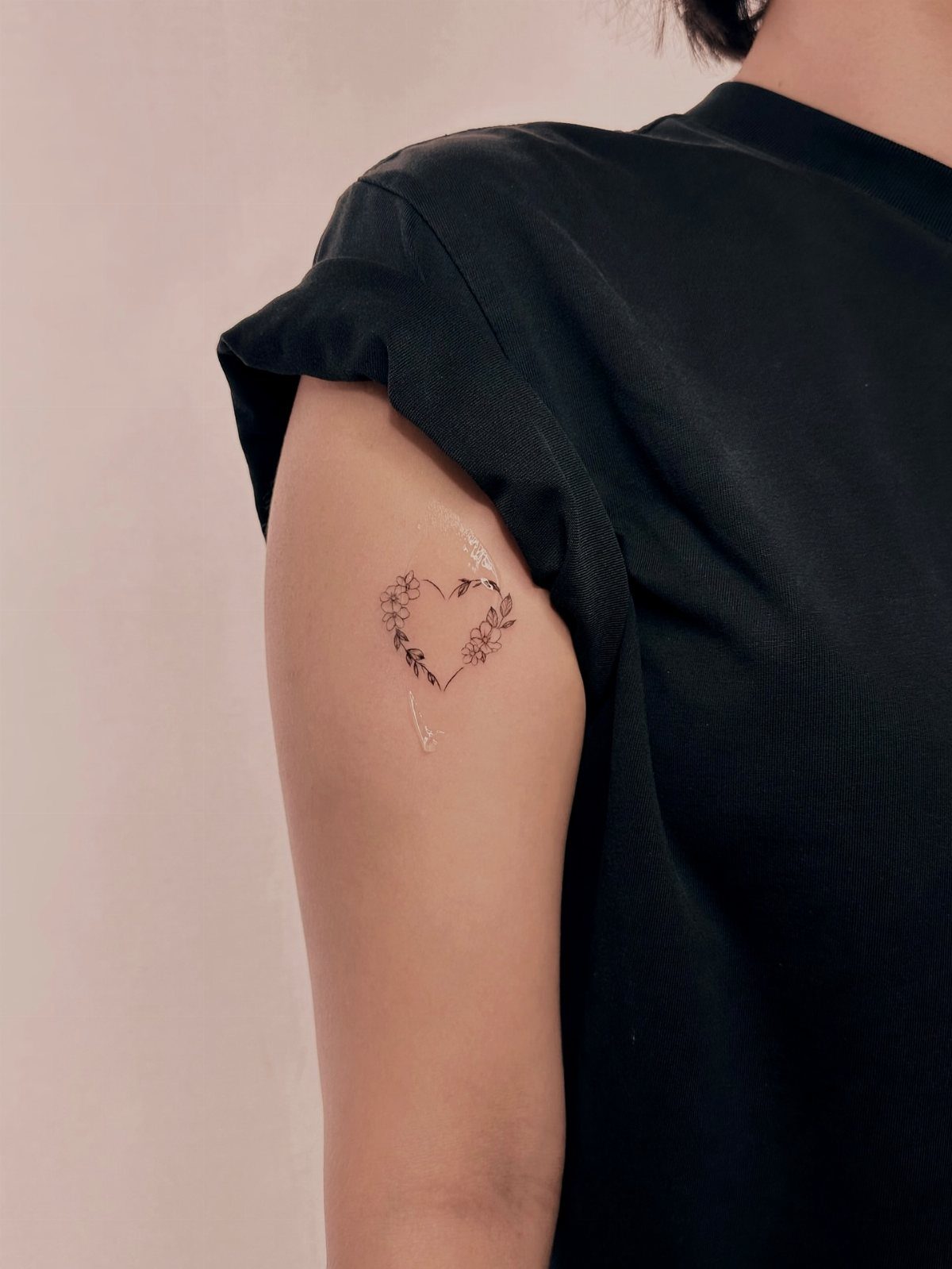 A small fine line tattoo of a heart surrounded by delicate flowers on an upper arm.