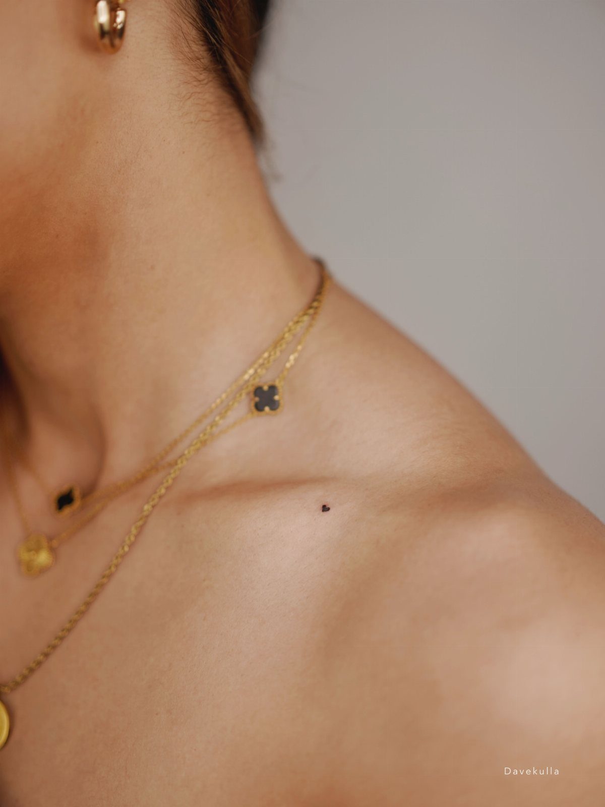 15 Tattoo Placement Heart Ideas for Meaning and Style 6 A tiny black heart tattoo placed on a person's collarbone near gold necklaces.