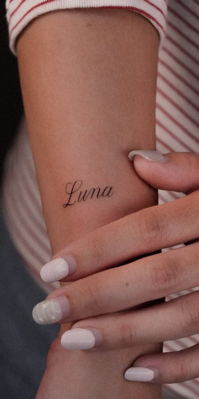 A small script tattoo reading Luna on a person's wrist with manicured nails.