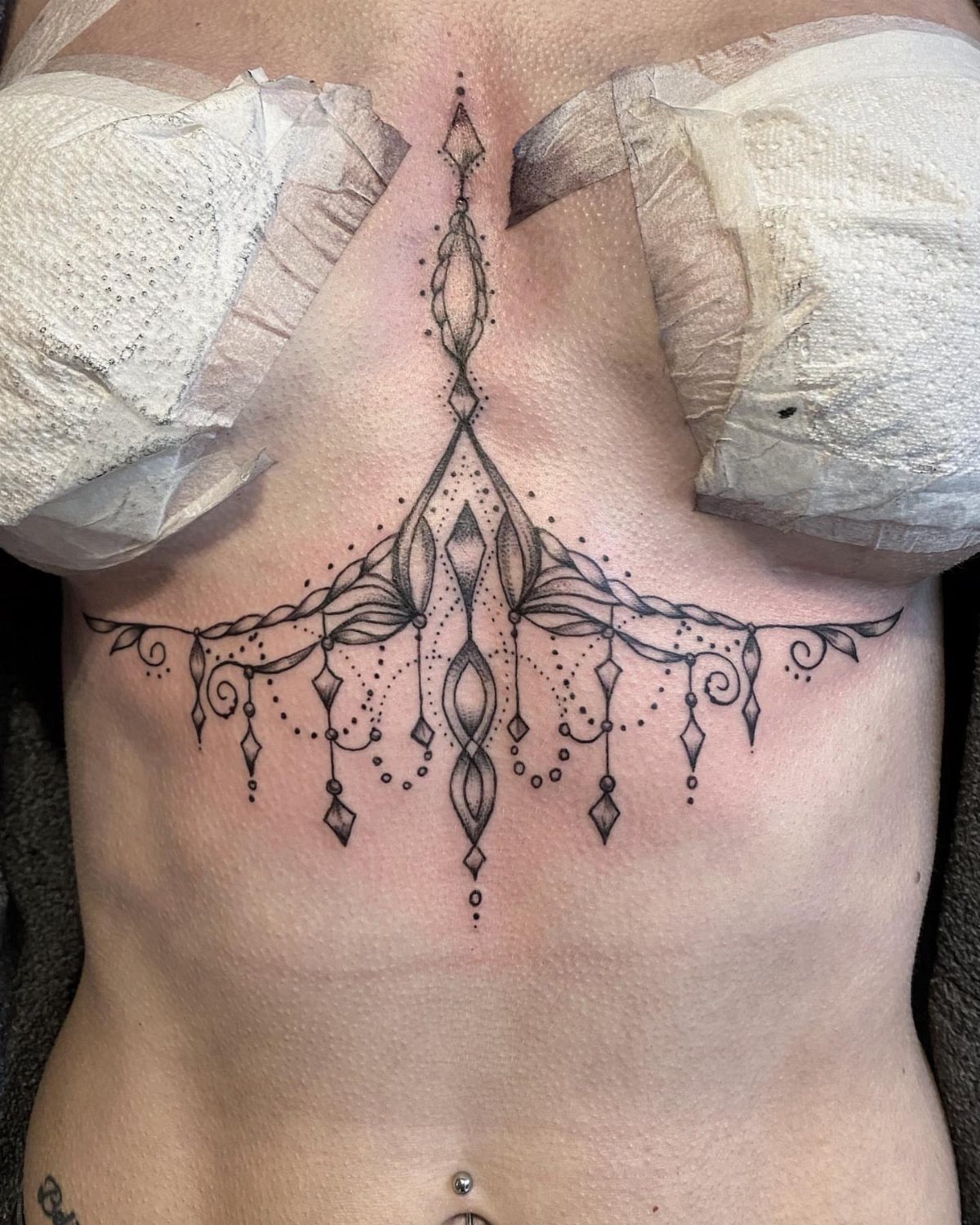 9 Chest Tattoo Ideas for Women: Floral, Sternum, Underboob & More 2026 4 An ornamental black ink tattoo follows the curve of a woman's underboob area.