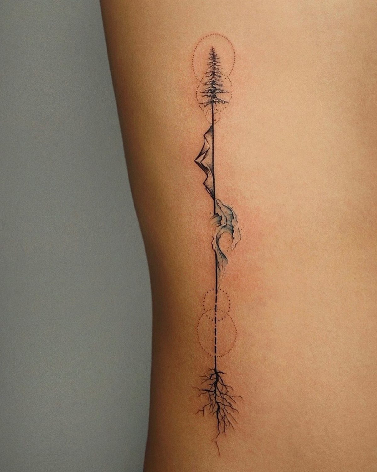 A vertical fine-line tattoo of a tree, mountains, and roots on a person's ribs.