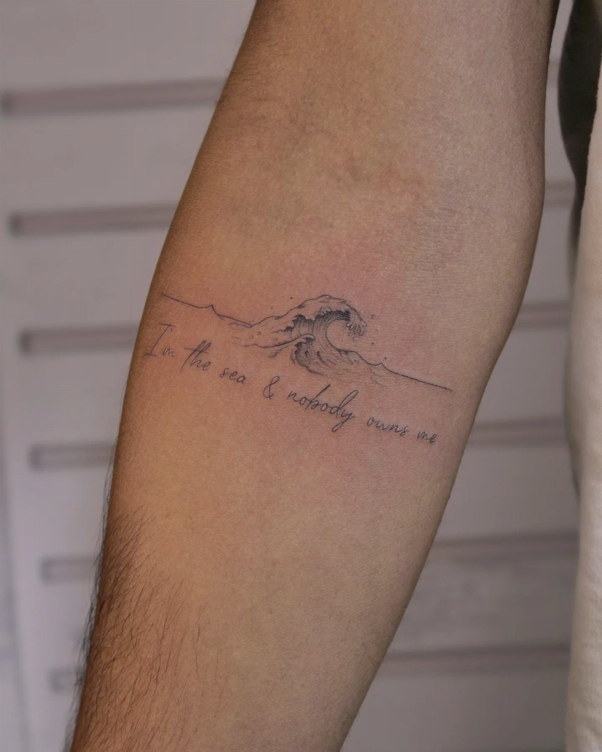 Fine Line Travel Tattoo: 10 Designs That Stay Light on Skin 9 A fine line tattoo of a crashing ocean wave with cursive text on a forearm.